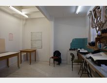 Ein schlichter Arbeitsraum mit weiß gestrichenen Wänden, einfachen Holztischen auf Rollen, einem Regal mit Malutensilien und blauen Kacheln an einer Teilwand. Im Vordergrund sind ein Stuhl und ein langer Arbeitstisch mit Papieren und Materialien zu sehen.