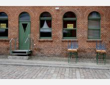 Fassade eines Backsteingebäudes mit grüner Eingangstür und drei Segmentbogenfenstern. Vor der Tür führt eine kleine Treppe mit Metallgeländern, daneben stehen draußen zwei zusammengeklappte Holzbänke auf Metallgestellen.