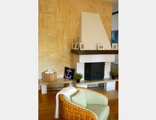 Wohnzimmer mit hellem Holzboden, einer gemusterten beigefarbenen Wand und einem weißen Kamin mit schwarzer Feuerstelle. Vor dem Kamin steht ein Sessel mit Korbrahmen, daneben Dekorationselemente wie Pflanzen und ein Kissen mit Anker-Motiv.