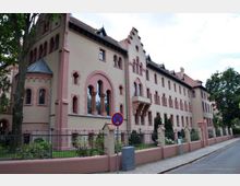 Historisches Gebäude mit rosa- und cremefarbenem Putz, Bogenfenstern und verzierten Giebeln im neugotischen Stil. Im Vordergrund ist ein Zaun mit Garten sowie ein Gehweg und eine schmale Straße sichtbar.