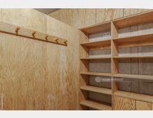 Raum mit Wänden und Einrichtung aus hellem Sperrholz, bestehend aus einer Wand mit Hakenleiste und einer Regalwand mit offenen Fächern. Im Hintergrund sind Teile einer Betonwand sichtbar.