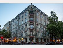 Eckgebäude aus der Gründerzeit mit dekorativer Klinkerfassade und Balkonen, gelegen an einer belebten Straßenkreuzung mit Geschäften, Restaurants und Fußgängern. Im Vordergrund stehen geparkte Fahrräder und Autos sowie Bäume und Straßenlaternen.