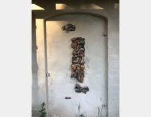 Relief an einer cremefarbenen Wand in einem eingelassenen, bogenförmigen Rahmen. Das Kunstwerk zeigt eine abstrakte, zusammengesetzte Figur und kleinere Elemente, darunter ein flaches Schild mit der Aufschrift „Juventud“ und niedrige Pflanzen am unteren Rand.