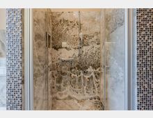 Moderne Dusche mit Glasabtrennung und Handbrause vor großformatigen, marmorierten Steinfliesen. Seitlich sind schmale Streifen aus kleinen Mosaikfliesen in Blau- und Brauntönen zu sehen.