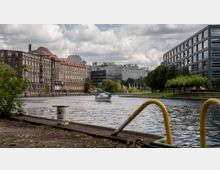 Ein Fluss mit einem kleinen Segelboot in einer urbanen Umgebung, umgeben von alten Backsteingebäuden auf der linken Seite und modernen Glas- und Bürogebäuden auf der rechten Seite. Im Vordergrund befinden sich Bodenelemente eines Kais sowie grüne Bäume entlang der Uferpromenade.