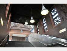 Innenansicht eines U-Bahnhofs mit Ziegelwänden, mehreren Lampen an der Decke und einem Metallgeländer entlang der Treppe. Große Fenster mit geometrischen Mustern lassen Licht in den Raum, während eine Rolltreppe und Stufen nach oben führen.