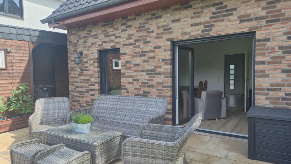Gepflasterte Terrasse an einem Haus mit Klinkerfassade und grauem Ziegeldach, ausgestattet mit einer Loungegarnitur aus grauem Rattan und Couchtisch. Rechts führt eine große, geöffnete Schiebetür in den Wohnbereich; links ist ein angrenzender Holzschuppen mit Wandlampe zu sehen.
