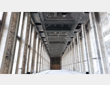 Ein langer, leerer Korridor in einem Gebäude mit hohen, symmetrisch angeordneten Marmorsäulen, teilweise freiliegenden Ziegelwänden und einer dunklen, rechteckig strukturierten Decke. Große Fenster entlang der linken Seite lassen Tageslicht in den Raum.