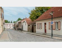 Gepflasterte, ruhige Straße mit historischen, einstöckigen Häusern mit roten Ziegeldächern und Holzklappläden. Rechts steht ein Schild mit Parkverbot, im Hintergrund sind Bäume und ein weiteres Gebäude sichtbar.