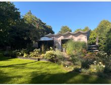 Modernes, flaches Gartenhaus mit markant geschwungenem Wellendach, umgeben von dichtem Baumbestand. Im Vordergrund liegen ein gepflegter Rasen, blühende Beete mit Ziergräsern und ein kleiner Treppenweg zur Terrasse mit Sonnenschirm.