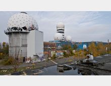 Ehemalige Abhörstation auf dem Teufelsberg in Berlin mit mehreren großen weißen Kuppeln auf industriellen Gebäuden, umgeben von überwachsender Vegetation und mit Graffiti bedeckten Wänden. Im Vordergrund ist eine asphaltierte Fläche mit Pfützen und metallischen Bauteilen zu sehen.