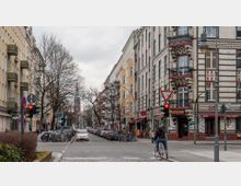 Straßenecke in einer urbanen Umgebung mit mehrstöckigen Wohnhäusern im Altbaustil; links hell verputzte Gebäude, rechts ein Gebäude mit dekorativer Klinkerfassade. Im Hintergrund ist die Spitze eines Kirchturms zu sehen, während die Straße mit geparkten Autos, Fahrradständern und Passanten belebt ist.