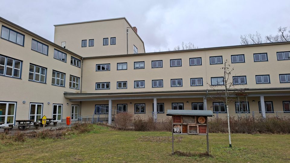 Mehrstöckiger, beige gestrichener Gebäudekomplex mit vielen rechteckigen Fenstern und einem überdachten Eingangsbereich an der Rückseite. Davor liegt eine Rasenfläche mit Sträuchern, einem kleinen Baum und einem Insektenhotel auf einem Gestell.
