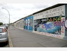 Teil der Berliner Mauer, bedeckt mit farbenfrohen Graffitis und Wandgemälden, darunter Texte und Gesichter. Rechts eine befahrene Straße mit parkenden Autos sowie modernen Straßenlaternen, im Hintergrund eine städtische Umgebung.