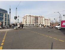 Straßenkreuzung in einer Stadt mit Fußgängern, einem Radfahrer und Fahrzeugen, darunter ein Lieferwagen mit Werbung. Im Hintergrund ein mehrstöckiges Wohngebäude mit Balkonen sowie moderne Bürogebäude auf der linken Seite.