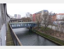 Fußgängerbrücke mit gläsernem Aufbau über einem Kanal, umgeben von Bäumen und mehrstöckigen Wohn- und Bürogebäuden. Links verläuft ein erhöhtes Bahngleis mit einem schmalen Holzsteg, im Hintergrund sind moderne und ältere Architektur erkennbar.