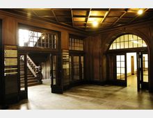 Historischer Innenraum mit hölzernen Wandverkleidungen und einem verzierten Kassettendeckenmuster; eine breite Holztreppe ist durch eine Türöffnung sichtbar, daneben Glas- und Holztüren mit großzügigen Fensterflächen. Schilder mit Informationen befinden sich an den Wänden.