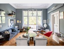 Wohnzimmer mit modernem Design, geprägt von hohen Decken mit Stuck, großen Fenstern und einer Glastür, die auf einen Balkon führt. Der Raum ist eingerichtet mit einem großen Sofa, verschiedenen Sesseln, einem runden Tisch und einer Hängelampe; die Wände sind grau-blau gestrichen und mit Bildern dekoriert.