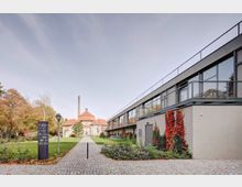 Moderner Gebäudekomplex mit großen Glasfenstern auf der rechten Seite und einem historischen Gebäude mit rotem Ziegeldach sowie einem hohen Schornstein im Hintergrund. Dazwischen verläuft ein gepflasterter Weg durch eine gepflegte Grünanlage mit Blumenbeeten, Rasenflächen und Bäumen.