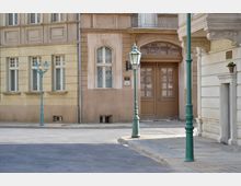 Straßenszene mit historischen Gebäudefassaden in Erdtönen, verzierten Fenstern und hölzernen Türen; im Vordergrund stehen grüne Straßenlaternen auf Steinpflaster.