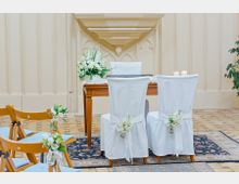 Ein hell dekorierter Raum für eine Trauung mit einem Holztisch, auf dem weiße Blumen und Kerzen stehen, und zwei mit weißen Hussen bedeckten Stühlen, die mit Blumengestecken verziert sind. Im Vordergrund sind Holzstühle mit blauen Sitzpolstern und Blumenschmuck, und im Hintergrund sind pastellfarbene, verzierte Wandpaneele zu sehen.