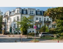 Mehrstöckiges Wohngebäude im Stil der Gründerzeit mit heller Fassade, Mansardendach und verzierten Balkonen, umgeben von Bäumen und niedrigem Grün. Im Vordergrund befinden sich Straßenschilder, ein parkendes Auto und ein Pflastersteinweg.