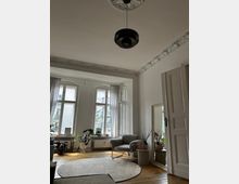 Helles Wohnzimmer mit hohen Decken und Stuckverzierungen, großen Sprossenfenstern und einem hölzernen Dielenboden. Ein grauer Sessel steht neben einem runden Teppich, umgeben von Pflanzen und einem großen Spiegel, vor dem helle Vorhänge hängen.