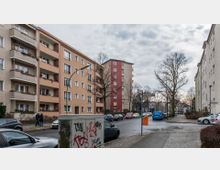 Straßenszene in einem Wohngebiet mit mehrstöckigen Wohnblöcken in beige, orange und roter Farbe, vereinzelt mit Balkonen. Am Straßenrand geparkte Autos, ein grauer Verteilerkasten mit Graffiti und kahle Bäume prägen das Bild, begleitet von Straßenschildern und Gehwegen.