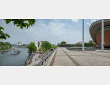 Weitläufiger Platz an der Spree mit Blick auf moderne Architektur, darunter das geschwungene Dach der Kongresshalle (Haus der Kulturen der Welt) rechts und ein Gebäude mit rundem Fenster links. Im Hintergrund ist ein Fernsehturm sichtbar, und auf dem Fluss fahren Ausflugsboote.