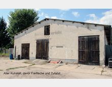 Ein alter Kuhstall aus hellem Putz mit Holzschiebetoren und leicht abgenutzten Fassaden, davor eine Freifläche mit Betonboden und Zufahrt; im Hintergrund Bäume und blauer Himmel.