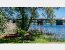 Flussufer mit Schilf und einem Baum im Vordergrund, dahinter eine Brücke mit fahrenden Lastwagen, die über den Fluss führt. Im Gras liegen zwei alte, umgestürzte Boote.