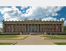 Frontale Ansicht des Alten Museums in Berlin, mit seiner klassizistischen Fassade, einer Reihe von Säulen, einem lateinischen Schriftzug auf dem Gesims und Skulpturen auf dem Dach. Im Vordergrund ein rechteckiger Platz mit Rasen, Gehwegen und Besuchern.