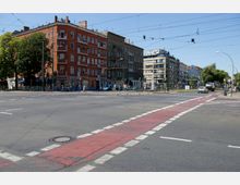 Straßenkreuzung in einer städtischen Umgebung mit mehrstöckigen Wohn- und Geschäftshäusern, darunter ein rotes Gebäude mit Balkonen und ein moderneres Glasgebäude im Hintergrund. Zu sehen sind Verkehrsmarkierungen, eine Fahrradspur sowie Fußgänger, Fahrräder und Autos.