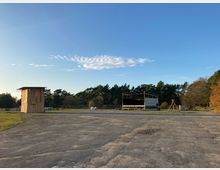 Weitläufiges Gelände mit einer asphaltierten Fläche im Vordergrund, einer Holzkonstruktion links und einer offenen Bühne rechts. Im Hintergrund sind Bäume sowie ein klarer blauer Himmel mit einzelnen Wolken zu sehen.