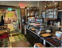 Gemütliches Café mit Theke auf der rechten Seite, auf der Speisen und Getränke wie Kuchen, Flaschen und Gläser präsentiert werden. Im Hintergrund sind ein Sitzbereich mit Holztischen, Stühlen und grünen Wandgemälden sowie ein retro-geometrischer Fliesenboden erkennbar.