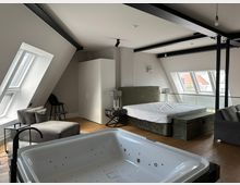 Modernes Dachgeschoss-Schlafzimmer mit schrägen Wänden, sichtbaren schwarzen Stahlträgern und großen Dachfenstern. Im Raum stehen ein Bett mit gepolstertem Kopfteil, ein Kleiderschrank, Sitzmöbel und im Vordergrund eine eingelassene Whirlpool-Badewanne auf Holzboden.