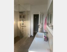 Modernes Badezimmer mit offener, verglaster Dusche auf der linken Seite, einem rechteckigen Waschbecken auf einer weißen Ablage rechts und einer geöffneten weißen Tür im Hintergrund. Wandfliesen mit einem Streifenmuster dekorieren den Duschbereich.