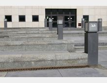 Außenbereich mit breiten Steinstufen, flankiert von grauen quadratischen Pfeilern mit Glaselementen, die als Laternen dienen. Im Hintergrund ist eine beige Fassade mit mehreren schwarzen Fenstern und einer großen Eingangstür erkennbar.