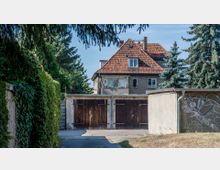 Zweistöckiges Wohnhaus mit rotem Ziegeldach und einer kleinen überdachten Terrasse, im Hintergrund von Bäumen umgeben. Im Vordergrund sind eine Reihe von Garagen mit Holztoren und eine mit Graffiti besprühte Mauer zu sehen.