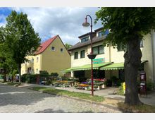 Eine kleine Blumenhandlung mit grün-weiß gestreifter Markise und vielfältigen Pflanzen auf dem Gehweg vor einem zweigeschossigen, gelben Gebäude. Im Vordergrund befinden sich Kopfsteinpflaster, ein Baum und eine Straßenlaterne mit Blumenampel.