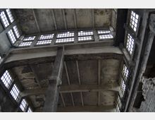 Innenansicht eines verlassenen Industriegebäudes mit Betonträgern, hohen Decken und rechteckigen Fenstern, die gleichmäßig verteilt sind. Die Wände und Decke zeigen deutliche Spuren von Verfall, darunter Abblätterungen und Verfärbungen.