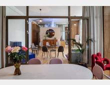 Moderner Innenbereich eines Veranstaltungs- oder Besprechungsraums mit Glaswänden und Holzboden. Im Vordergrund stehen ein runder Tisch mit Blumen und rosa Stühlen, im Hintergrund befinden sich graue Stühle im Wartebereich, ein runder Spiegel, eine Holzkommode und eine Neonbeleuchtung mit dem Text „Now? YES!“.