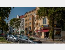 Straßenszene in einer städtischen Umgebung mit mehrstöckigen Wohngebäuden und Geschäften im Erdgeschoss, darunter eine Bäckerei mit rotem Sonnendach. Vor den Gebäuden sind Autos geparkt, und Bäume säumen den Straßenrand.