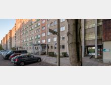 Mehrstöckiger Plattenbau in einer Wohnstraße mit geparkten Autos im Vordergrund und einem Straßenschild mit der Aufschrift „Pablo-Neruda-Block“; rechts ein Eingang mit Rampe und einem Schild, das auf eine Arztpraxis hinweist.