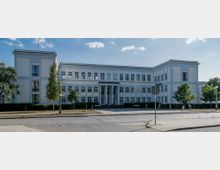 Klassizistisches, weißes Gebäude mit symmetrischem Baukörper und einem Mittelrisalit mit Säulen am Eingangsbereich. Umgeben von Bäumen, Rasenflächen und einer gepflasterten Straße im Vordergrund.