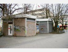 Zwei kleine, rechteckige Betonpavillons mit grauen Fassaden und Flachdächern, bedeckt mit Graffiti und Plakatanschlägen, stehen nebeneinander auf einem gepflasterten Platz. Im Hintergrund sind kahle Bäume und ein größeres Gebäude sowie Geschäfte sichtbar.
