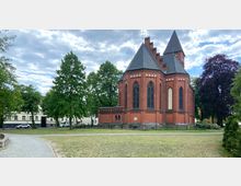 Backsteinkirche mit gotischen Spitzbogenfenstern und schwarzen Schieferdächern, umgeben von Rasenflächen, Bäumen und einem gepflasterten Weg; im Hintergrund stehen Wohngebäude.