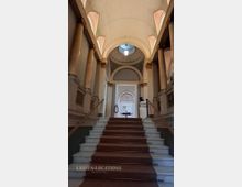 Blick in ein klassizistisches Treppenhaus mit breiter Marmortreppe und mittigem Teppichläufer, flankiert von Säulenreihen und Balustraden. Über den Treppen wölben sich hohe Rundbögen und eine Decke mit Oberlichtkuppel sowie hängender Leuchte.