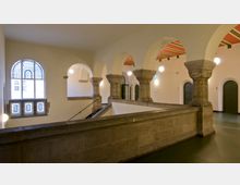 Offener Innenbereich eines historischen Gebäudes mit gewölbten Steinsäulen, einem Geländer aus Stein und großen Rundbogenfenstern mit dekorativen Glasornamenten. Die Decke ist mit Streifen in Rot und Grün gestaltet; runde Lampen hängen herab, und es sind mehrere geschlossene Türen sichtbar.