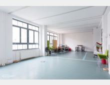 Ein heller Mehrzweckraum mit großen Fenstern und grauen Fensterrahmen, die viel Tageslicht hereinlassen. Der Raum hat eine minimalistische Einrichtung mit gestapelten Stühlen, Tischen, einem Pflanzenarrangement und einer weißen Wand als Hintergrund.